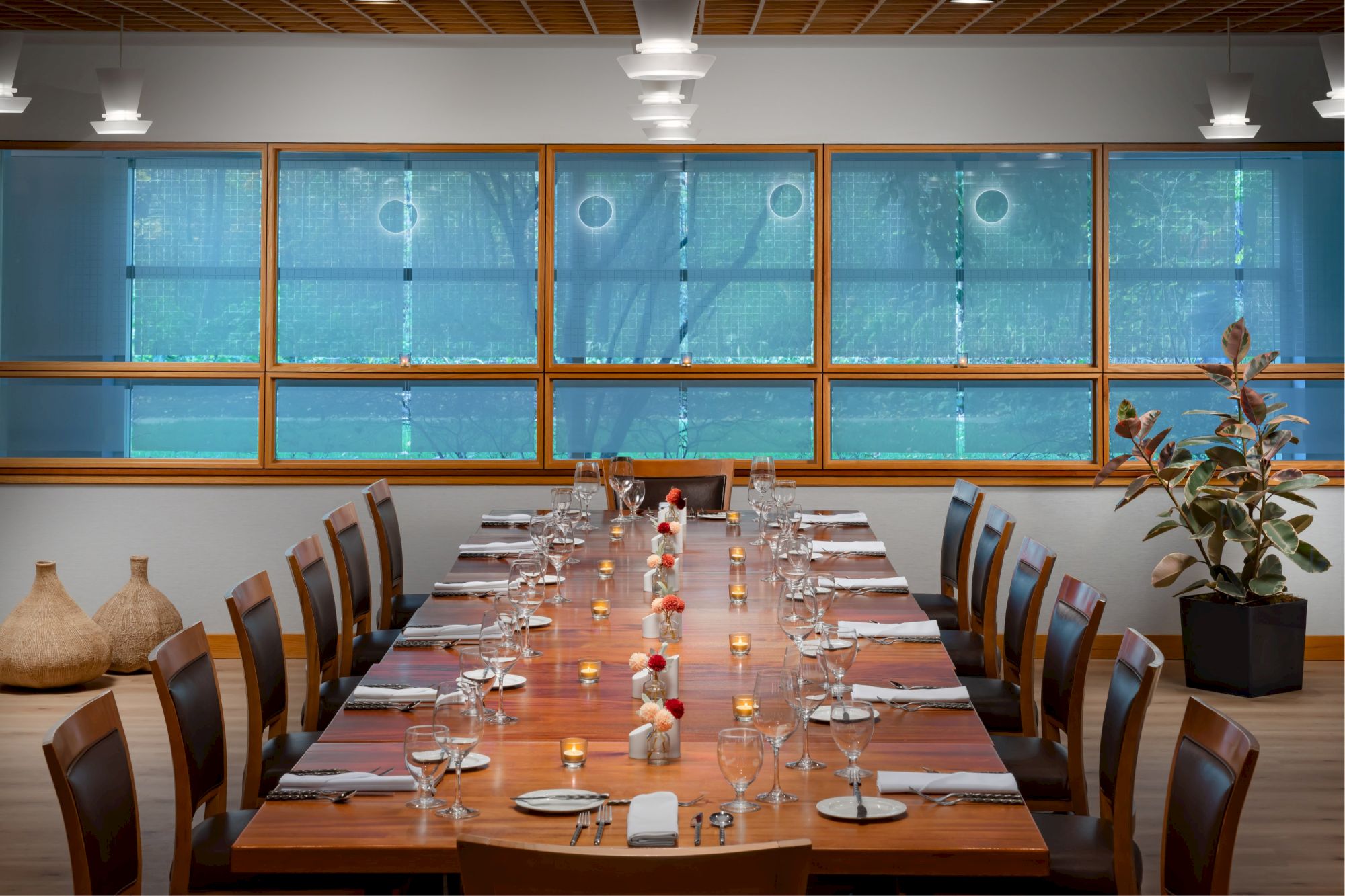 A long wooden dining table set for many guests in a bright, modern room with blue-tinted windows and a plant in the corner, ready for dinner.