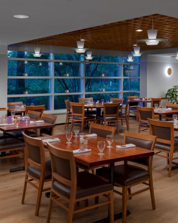 A modern dining area with wooden tables and chairs, wine glasses, pendant lighting, potted plants, and large windows, tidy and inviting.