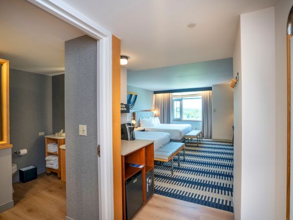 A hotel room suite with a separate bathroom, entry hall, desk, sofa, bed, and blue patterned carpet, plus a window view and light-colored walls.