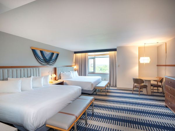 A bright hotel suite with two queen beds, a blue-and-white striped carpet, a bench at the foot of the bed, a desk area, and a window with curtains, all in soft neutrals.