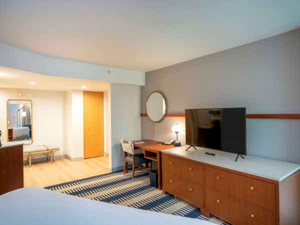 A modern hotel room with a large TV on a wooden dresser, a round mirror, a desk, and a patterned rug, plus a seating area and soft lighting.