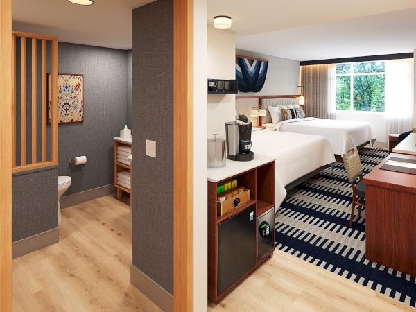 A hotel room with two beds, a desk, and a small kitchenette area; a separate entry area with a bench, and a blue-accent rug.