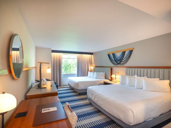 A bright hotel room with two large beds, a desk, round mirror, lamps, and a window with drapes featuring a blue accent, yielding a clean, modern vibe.