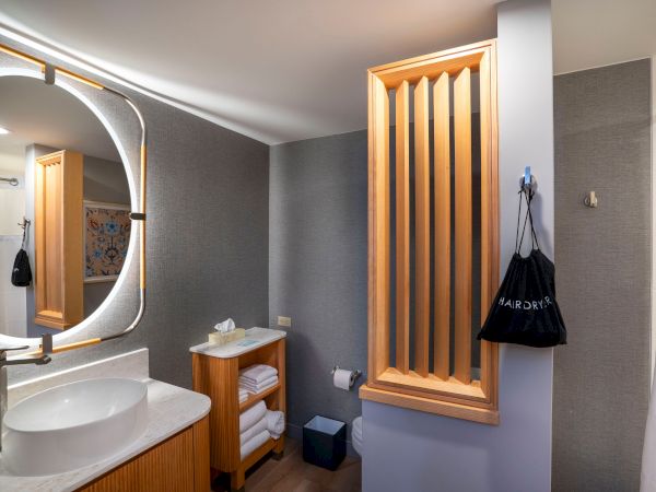 A modern bathroom with a round mirror, white sink, wooden cabinet, open shelf, gray walls, and a vertical wooden slat room divider, plus a hanging bag.