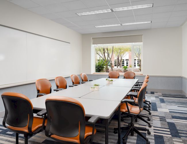 A modern conference room with a long table, orange-and-black chairs, whiteboards, and a window view of trees outside, ready for a meeting.