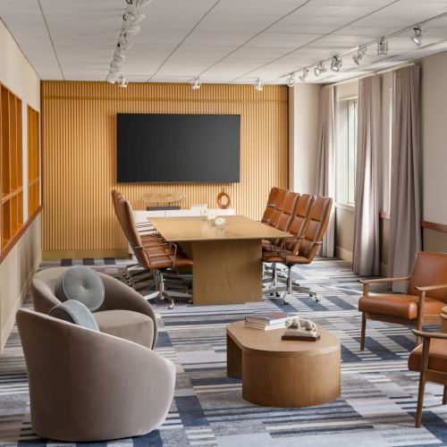 A modern conference room with a long wooden table, tan leather chairs, a large black screen on a wooden paneled wall, and a cozy seating area by windows.
