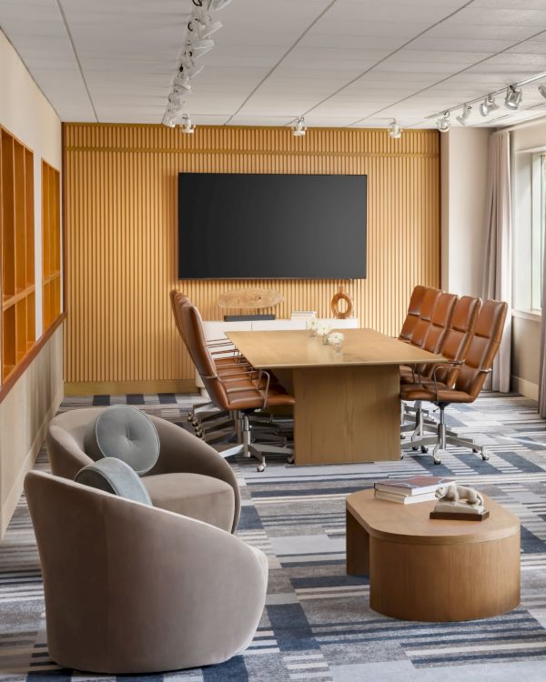 A modern conference room with a long wooden table, tan leather chairs, a large black screen on a wooden paneled wall, and a cozy seating area by windows.