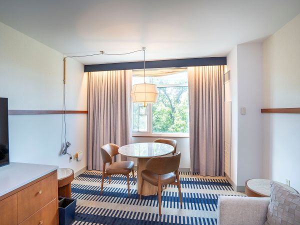 A bright hotel room with a round table and four chairs by a window, blue striped carpet, wall-mounted shelves, a TV on a wooden cabinet, and beige seating.