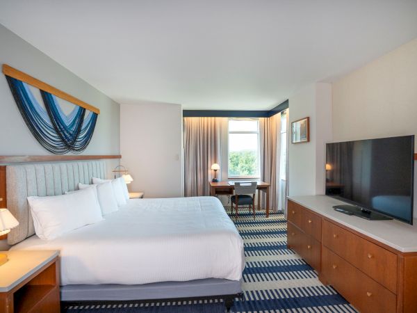 A hotel room with a king bed, nightstands, a wall art piece, a large dresser with a flat-screen TV, and a seating area by a window.