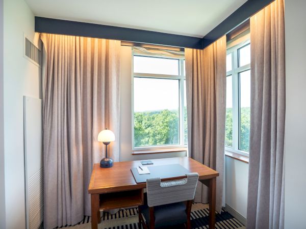 A small home office with a wooden desk, gray chair, table lamp, and large windows with beige curtains, overlooking greenery. Top it at 140 characters, always ending the sentence.