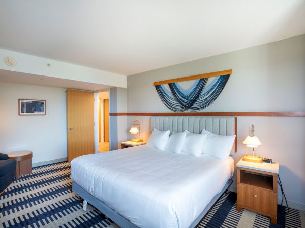 A bright hotel room with a large bed, two nightstands, lamps, a desk, and a doorway; blue and white striped carpet and a curved wall art above the headboard.