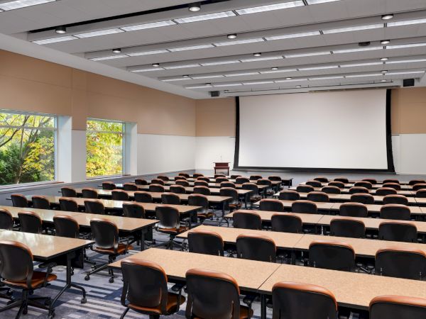 A large empty lecture hall with rows of desks and chairs facing a projection screen, windows on the side letting in natural light, and a podium at the front.