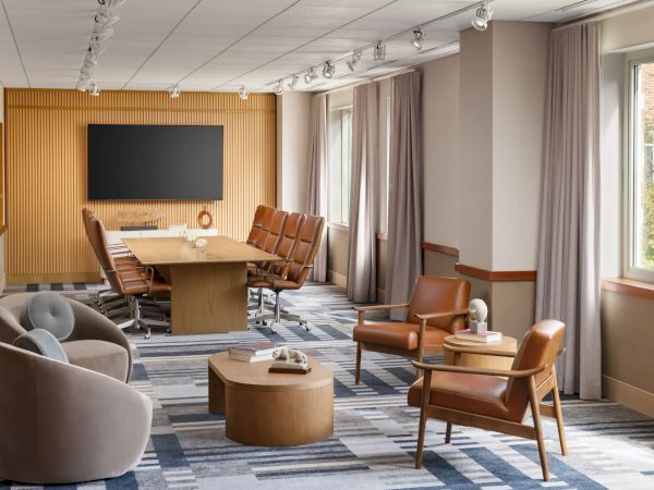 A modern conference room with a long wooden table, tan chairs, a wall-mounted screen, and a cozy seating area by the windows, stylish and bright.