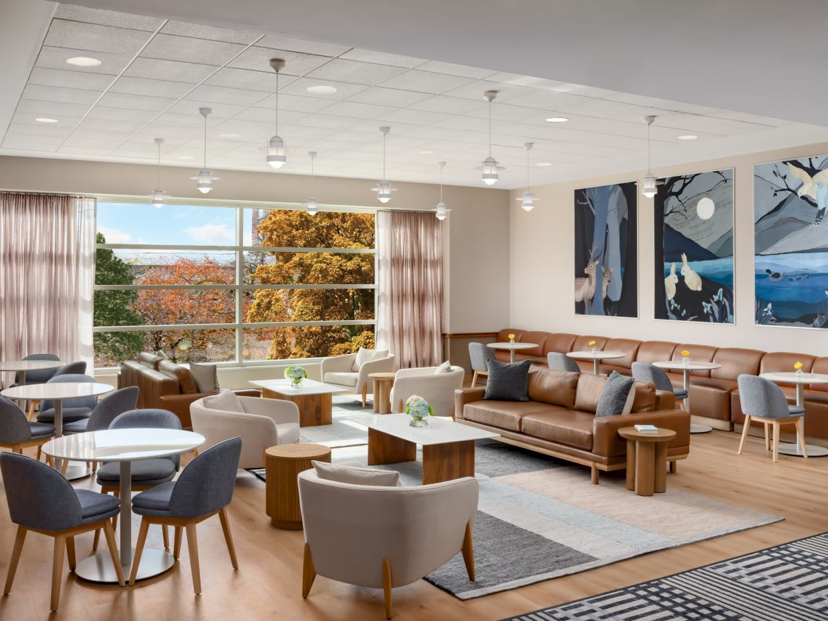 A bright, modern lounge with multiple seating areas, comfy sofas and chairs around round tables, large windows, and nature art on the wall.