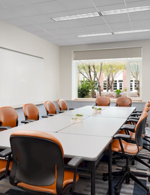 A modern conference room with a long white table, orange chairs, a whiteboard, and large windows letting in natural light.