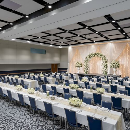 A spacious event hall set for a wedding or banquet, with long tables, white linens, blue chairs, floral centerpieces, and a decorated altar backdrop.