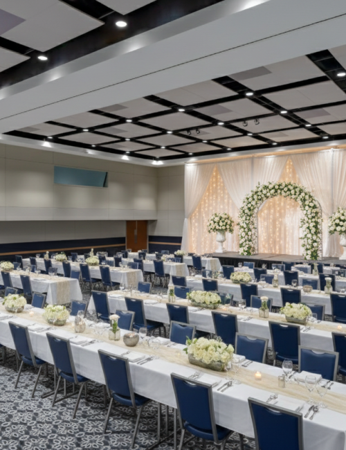 A large banquet hall set for a wedding or formal event with long head tables and rows of seating, floral arrangements, and a decorated stage with drapery.