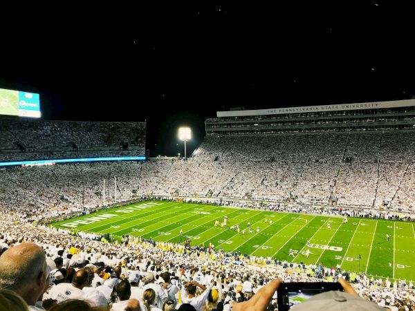 A packed night football stadium filled with cheering fans, white attire, bright lights, and a green field under the scoreboard glow.