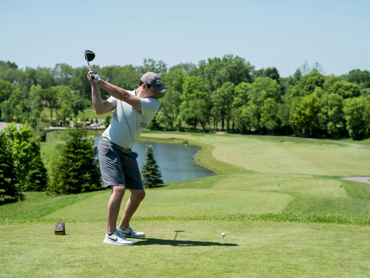 A man in a gray tank top and shorts swings a golf club on a sunny course, teeing off with trees and a water hazard in the background.