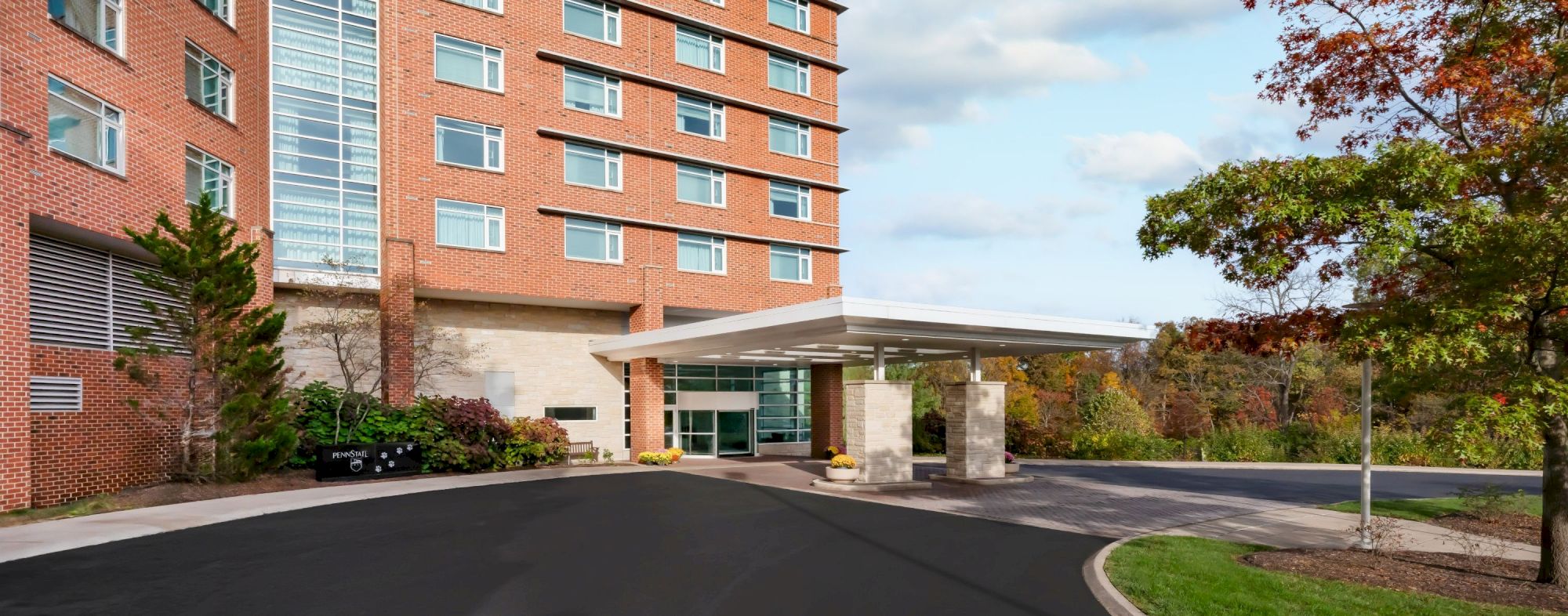 A brick hotel or apartment building with a glass atrium, driveway, and small trees; blue sky with scattered clouds, autumn foliage in the background.
