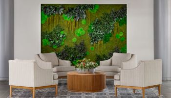 A modern living room with beige couches arranged around a round wooden coffee table, a greenery wall art focal point behind, and a patterned rug.