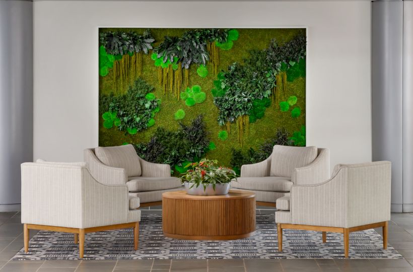 A modern living room with beige couches arranged around a round wooden coffee table, a greenery wall art focal point behind, and a patterned rug.