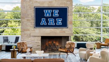 A modern living room with a stone fireplace, large windows, and a bold blue &ldquo;WE ARE&rdquo; sign above the mantel, seating surrounds the cozy hearth.