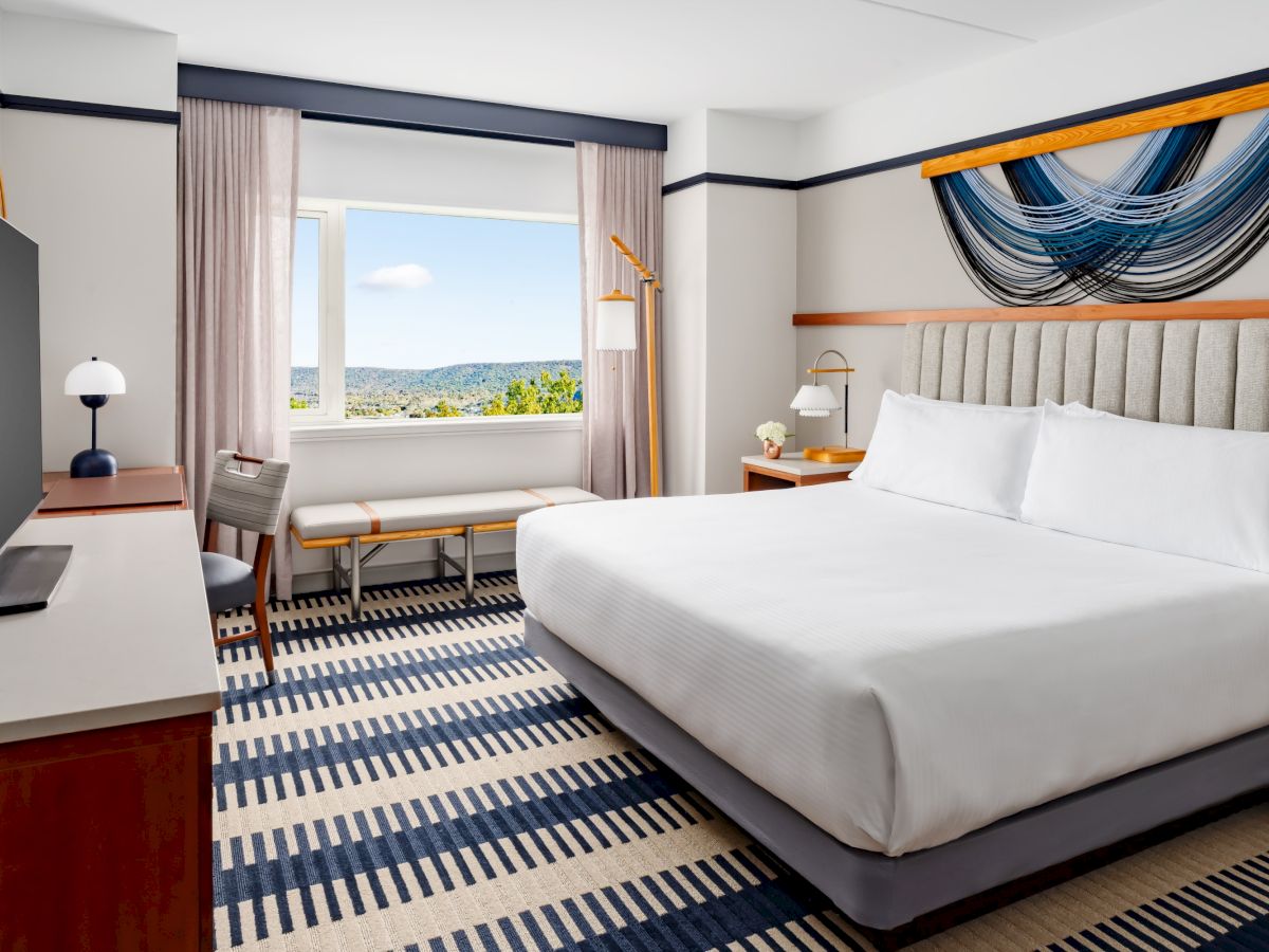 A bright hotel room with a king bed, striped carpet, desk by a window, seating area, and blue-gray drapery; a scenic view beyond the window. End.