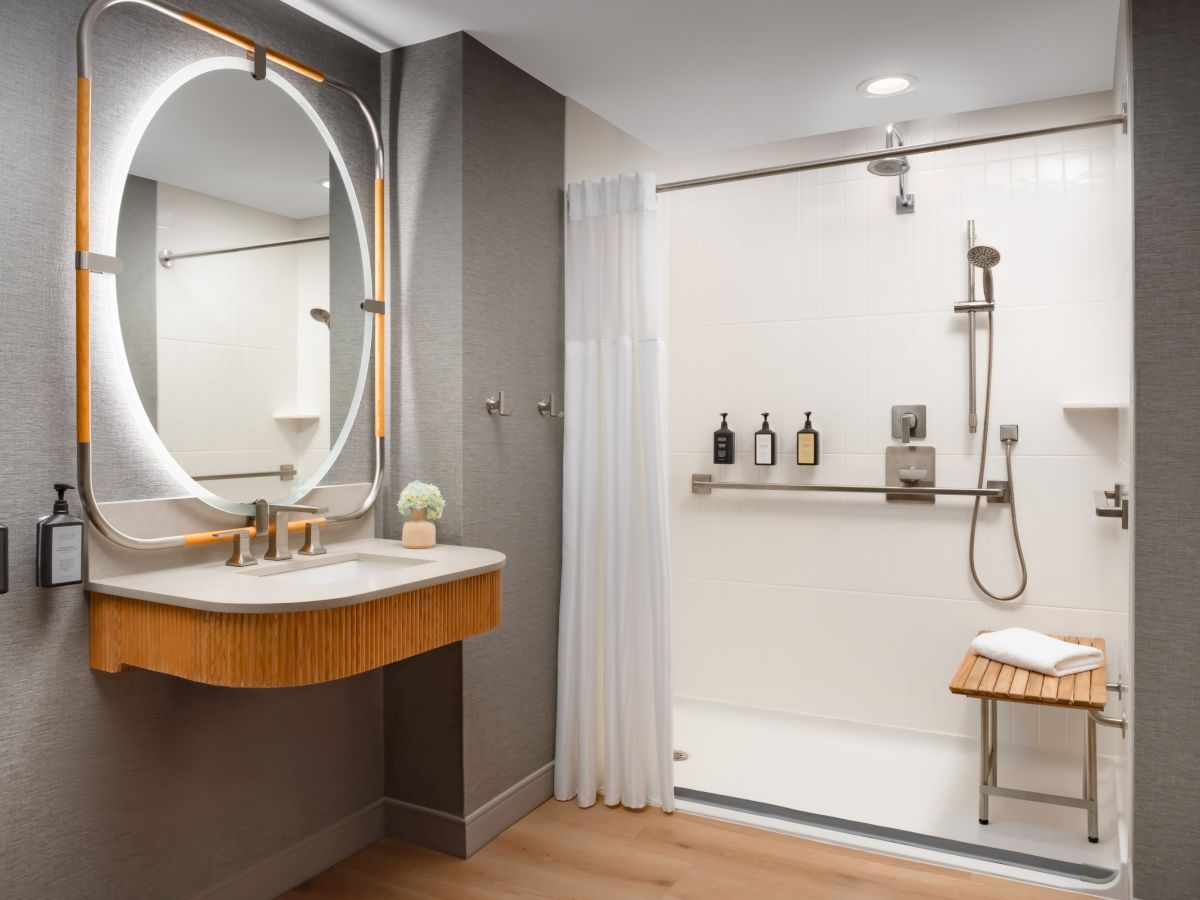 A modern bathroom with a round backlit mirror, wooden sink cabinet, gray walls, and a glass-enclosed shower featuring a rain shower and bench, plus toiletries and a towel.