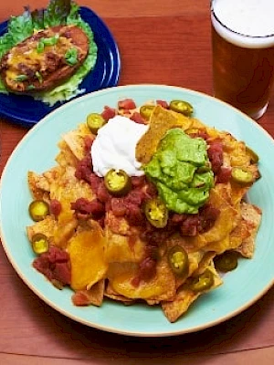Tortilla chips nachos with melted cheese, jalapeños, sour cream, plus a side of tacos or tostadas and a glass of beer on a wooden table.