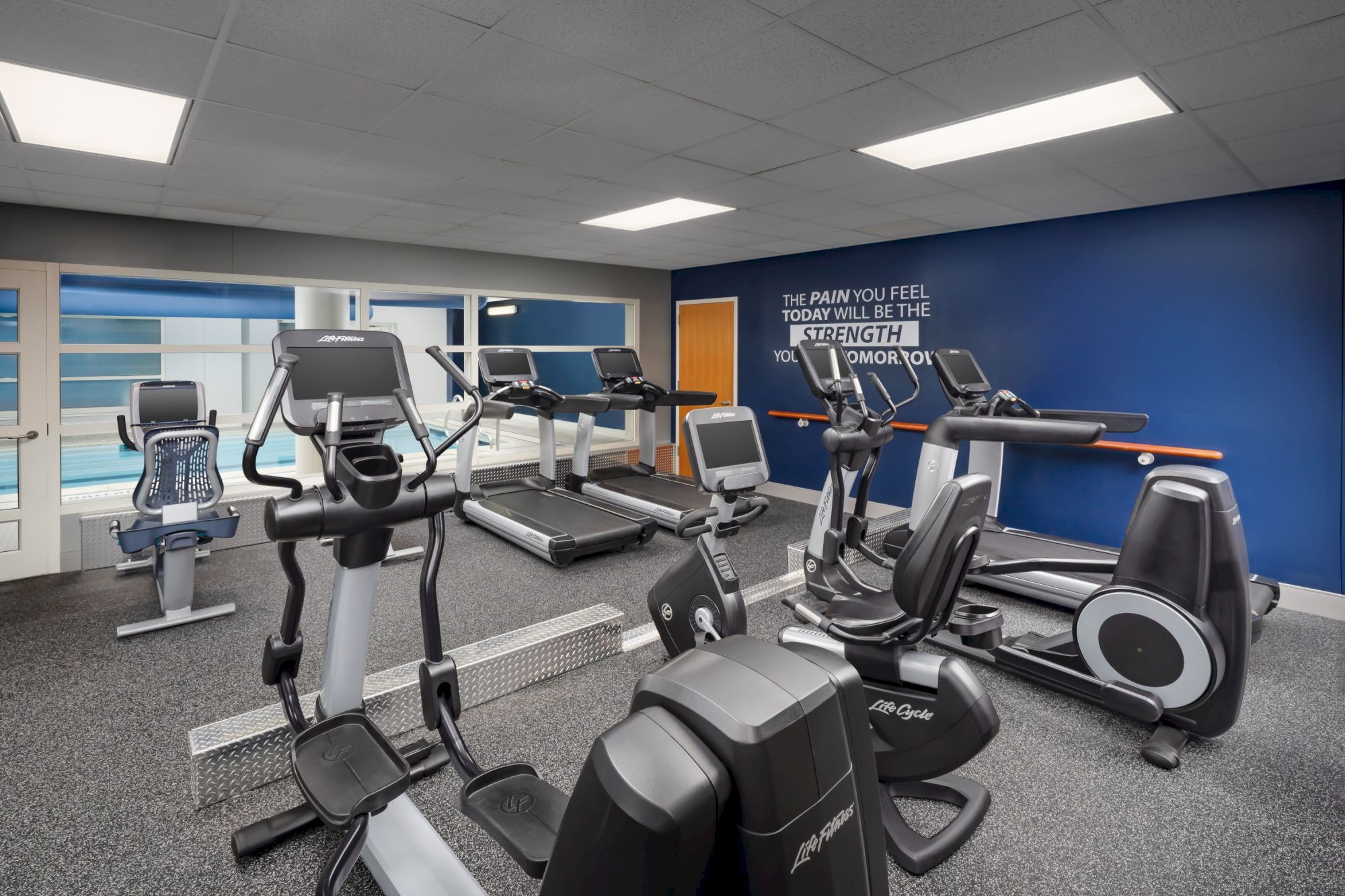 A bright hotel gym with treadmills, ellipticals, and bikes; blue accent wall, large windows, and motivational quote on the wall.